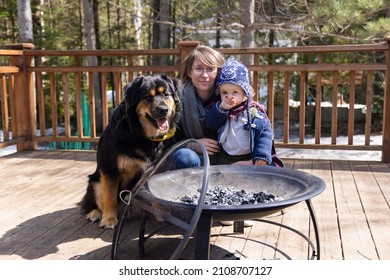 Family Portrait By A Metal Fire Pit In The Back Yard. Mother Poses With Shy Three Year Old Son And Large Burmese Cross Dog Wearing Winter Clothes.