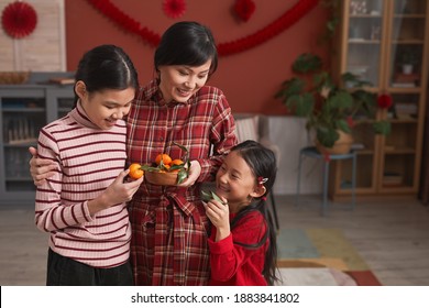 Family Portrait Of Chinese Woman And Two Lovely Daughters Standing Together In Living Room Taking Tangerines For Lunar New Year