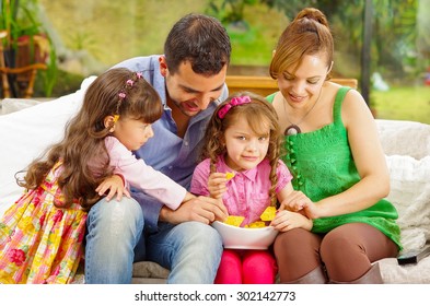 Family Portrait Of Father, Mother And Two Daughters Sitting Together In Sofa Sharing Bowl With Nachos On Little Girls Lap .