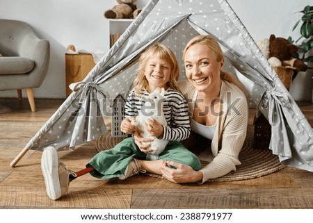 family portrait of happy girl with prosthetic leg holding soft toy near mother in play tent