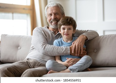 Family Portrait Happy Mature Grandfather Hugging Adorable Grandson, Smiling Older Man With Cute Little Boy Grandchild Looking At Camera, Sitting On Cozy Couch At Home, Two Generations Concept