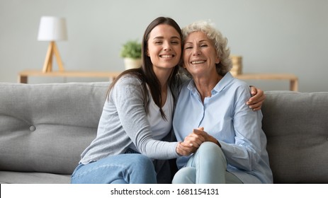 Family Portrait Of Happy Mature Mom Hug Cuddle With Grown-up Adult Daughter Sit Together On Couch In Living Room, Smiling Elderly Mother And Millennial Girl Child Embrace Posing For Picture At Home