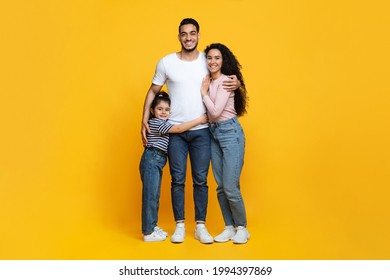 Family Portrait. Happy Middle Eastern Parents And Their Little Daughter Embracing Over Yellow Background, Full Length Shot Of Cheerful Arab Mother, Father And Child Hugging Together In Studio