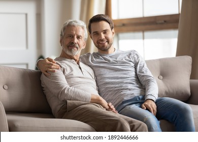 Family Portrait Of Happy Millennial Caucasian Man With Elderly 70s Father Sit Relax On Couch At Home. Smiling Mature Dad With Adult Grownup Son Rest On Sofa, Enjoy Weekend Together. Bonding Concept.