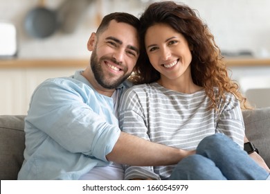 Family portrait of happy millennial husband and wife sit on couch hug cuddle look at camera posing, smiling young couple embrace show love affection, relax at home on weekend together