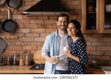 Family portrait happy young couple drinking coffee or tea in morning, standing in cozy kitchen at home, smiling wife and husband hugging, looking at camera, holding mugs, starting new day together