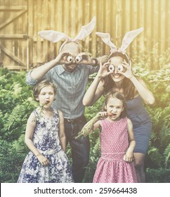 Family Portrait On Easter Parents Wearing Bunny Ears And Holding Up Silly Eyes Made From Eggs As Their Children Pose Eating Carrots Outside In The Spring Season.  Filtered For A Retro, Vintage Look.