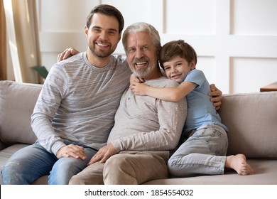 Family Portrait Smiling Mature Grandfather, Young Father And Little Boy Hugging, Sitting On Couch At Home, Happy Three Generations Of Men Looking At Camera, Posing For Photo In Living Room