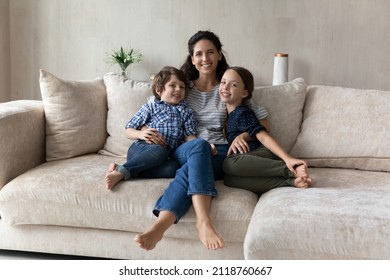 Family Portrait Smiling Mother Hugging Little Son And Daughter, Sitting On Cozy Couch At Home, Happy Loving Hispanic Young Mom Embracing Adorable Kids, Looking At Camera, Posing For Photo Together