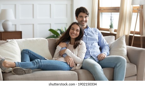 Family Portrait Smiling Woman And Man Hugging, Sitting On Cozy Couch In Living Room At Home, Happy Young Couple Posing For Photo, Loving Wife And Husband Cuddling, Looking At Camera Together