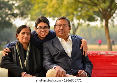 Family Portrait Of A Young Indian Woman With Her Mother And Father Sitting On A Red Bench, Posing In A Park In Delhi, India