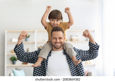 Family power. Strong young man carrying his little son on shoulders, both demonstrating biceps and smiling to camera at home, sport lovers concept