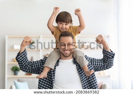 Family power. Strong young man carrying his little son on shoulders, both demonstrating biceps and smiling to camera at home, sport lovers concept