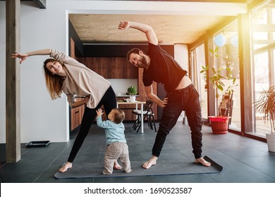 The Family Practices Yoga, Family Life In Isolation. Mom And Dad Are Doing Exercises, And Infant Child Creeps Next To His Parents. Sunset Light From The Windows. Family Quarantine