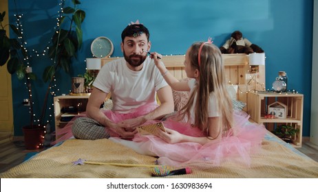 Family preparation for birthday fancy party. Little girl fairy applying makeup to her father. Portrait of silly happy young man smiling and sitting on bed. Two pretty magic fairies.
