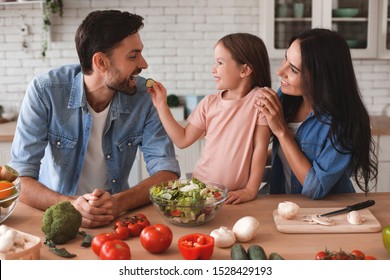 family preparing salad for lunch, daughter feeding father with slice of cucumber on the kitchen