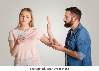 Family Problems, Indifference And Quarrel. Busy Young Wife Looking At Phone, Typing Message And Making Stop Hand Gesture For Husband, Want To Talk Isolated On Gray Background, Free Space, Studio Shot