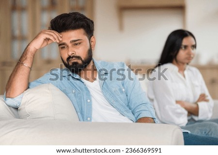 Family problems. Unhappy indian man sitting separate from discontented wife on sofa at home, couple ignoring each other, selective focus on thoughtful hindu guy