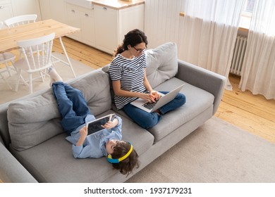 Family, quarantine, distance job concept. Happy mother freelancer remote work from home office on laptop during lockdown, sitting on sofa, little child playing on digital tablet and wear headphones. 