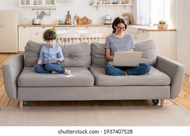 Family, quarantine, distance job concept. Happy mother freelancer remote work from home office on laptop during lockdown, sitting on couch, little son playing on digital tablet and wear headphones.  - Powered by Shutterstock