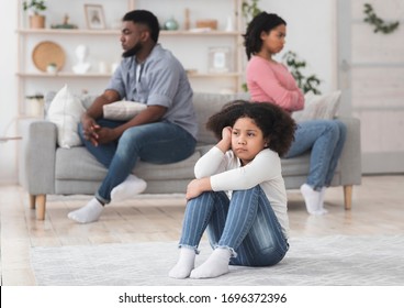 Family Quarrels Concept. Upset Little Black Girl Sitting On Floor Separately From Parents After Their Argue, Selective Focus On Kid