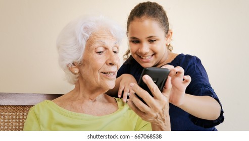 Family Relationship With Grandmother And Granddaughter. Happy Old Woman And Little Girl Using Smartphone, With Grandchild Helping Grandma.