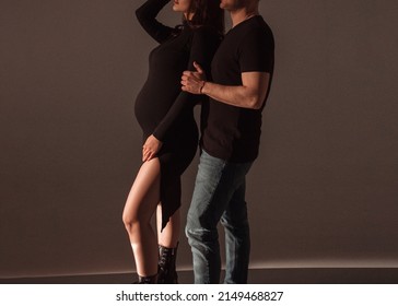 Family Relationship. Pregnant Couple. Man In Black T-shirt And Jeans Hugs Pregnant Girl In Black Slim Dress On A Dark Studio Wall Background. Pregnant Concept, Free Space