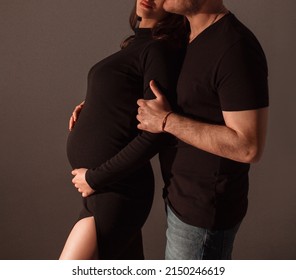 Family Relationship. Pregnant Couple. Man In Black T-shirt And Jeans Hugs Pregnant Girl In Black Slim Dress On A Dark Studio Wall Background. Pregnant Concept, Free Space