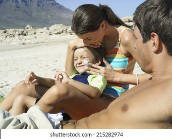 Family Relaxing On Beach, Boy (4-6) Sitting In Mother's Lap, Woman Applying Suncream To Son's Face, Side View