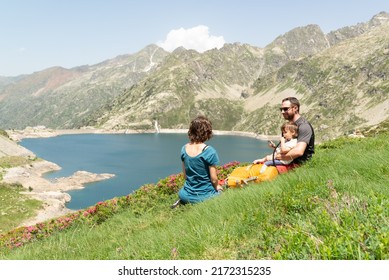 Family Resting On A Grassy Slope, Looking At Each Other And Drinking Water From Their Hydration Backpack With A Scenic Aerial Views Of A Lake Surrounded By Mountains.