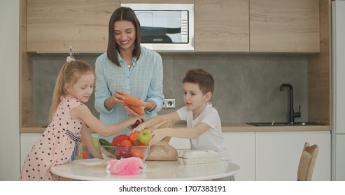 Family Returning Home From Shopping Trip Using Paper Bag Unpacking Groceries In Kitchen. Mother And Kids At Home.