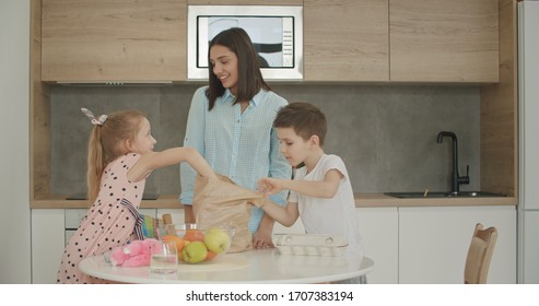 Family Returning Home From Shopping Trip Using Paper Bag Unpacking Groceries In Kitchen. Mother And Kids At Home.