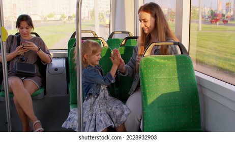 Family rides in public transport tram, mother with blonde girl sit together and playing game clapping hands, talking, laughing. Woman passenger with her daughter having fun in bus. Commuters. Train