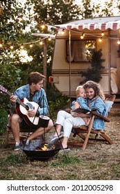 Family Rv Camping, Sit At Camp Fire With Guitar And Small Son Relax Outdoor Modern Caravan On Summer Road Trip. Happy Parents Enjoy Travel In Nature On Motorhome With Little Kid. Mobile Home Voyage