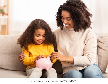 Family Savings. Cheerful Afro Mother Teaching Little Daughter How To Save Money Holding Piggybank Sitting On Sofa At Home