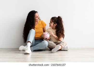 Family Savings. Happy Little Girl And Her Mom Holding Piggybank And Smiling While Relaxing On Floor Near White Wall At Home, Young Middle Eastern Mother Teaching Daughter Economy, Copy Space