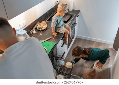 Una escena familiar en una cocina moderna. Una mujer carga el lavavajillas, un hombre ayuda, y un niño pequeño se sienta en el mostrador observando. Representa la vida diaria, el trabajo en equipo y las tareas domésticas en casa.