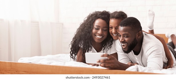 Family selfie. Cheerful african american father taking photo with his wife and daughter, relaxing in bed together - Powered by Shutterstock