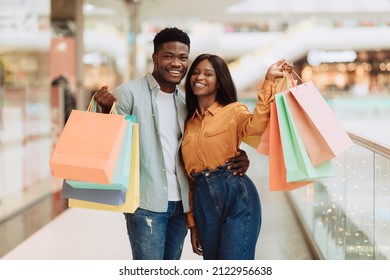 Family Shopping Concept. Portrait of two excited African American consumers standing in shopping mall, holding and showing colorful shopper bags, guy hugging lady enjoying their new purchase