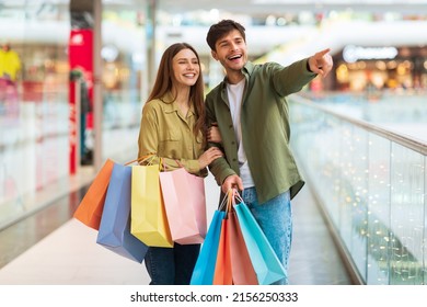 Family Shopping. Happy Spouses Holding Shopper Bags Pointing Finger Looking Aside Standing In Modern Hypermarket Indoor. Couple Advertising Sales Offer. Consumerism Concept - Shutterstock ID 2156250333