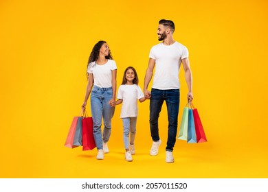 Family Shopping. Middle-Eastern Parents And Daughter Carrying Colorful Shopper Bags Walking Together Holding Hands Posing Over Yellow Background In Studio. Full Length