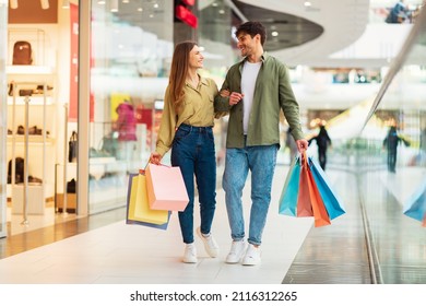 Family Shopping. Smiling Couple Carrying Colorful Shopper Bags During Great Sales Walking Holding Hands Buying New Clothes In Modern Hypermarket. Contented Customers In Mall Concept