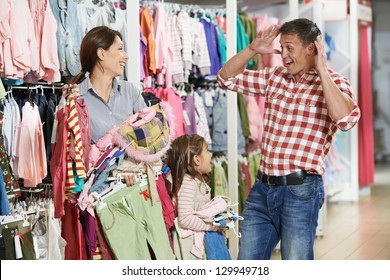 Family Shopping. Woman And Girl Holding Clothes With Highly Surprised Father Man During Shopping At Outerwear Supermarket