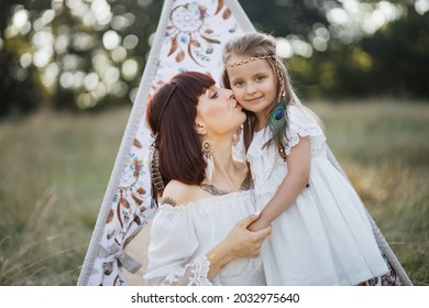 Family Shot In Native American Indin Style. Happy Young Mother Cuddles And Kisses Her Daughter In The Summer On Field, Sitting Near Boho Tee Pee Tent. Mom And Daughter Wearing White Dress And Feathers