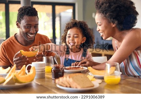 Family Shot With Parents And Daughter At Home Having Breakfast Spreading Jam On Bread At Table
