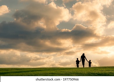 Family Silhouette Against A Bright Cloudy Sky At Sunset. Mother And Two Boys Walking Holding Hands