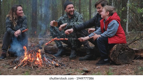 Family Sits Around A Campfire On A Summer Evening. Children With Parents Are Resting In The Woods. Family Frying Sausages Over A Fire In The Woods. Weekend In Nature In Good Company. 