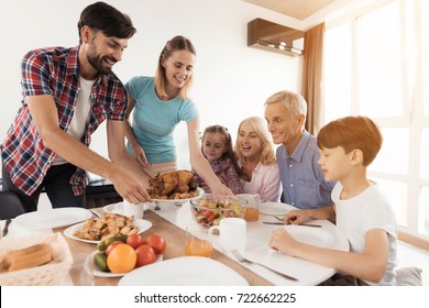 The Family Sits Down For Dinner On Thanksgiving. The Man Serves A Festive Turkey, His Wife Stands Beside Him And Serves A Salad, The Rest Sit And Look Forward To
