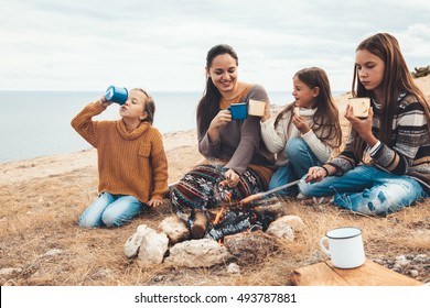 Family Sitting By The Fire And Frying Sausages In Field. Autumn Hike In Cold Weather. Warming And Cooking Near Flame Together.