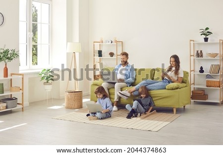 Family sitting in cozy spacious Scandinavian livingroom with wireless Internet connection at home, kids watching video on laptop and tablet, parents using their own devices. People, technology concept